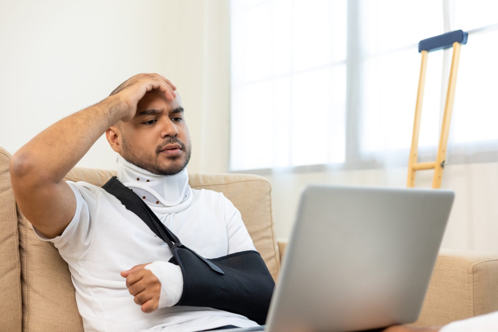 An injured man is distressed while looking at his computer. His arm is in a sling and there is a crutch next to him against the wall.