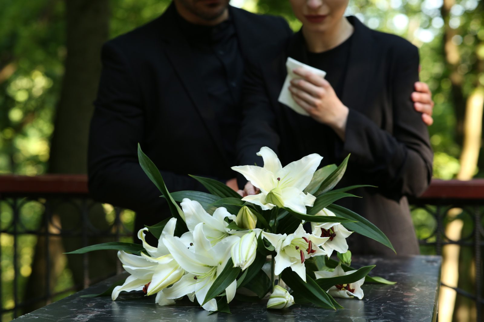 A grieving couple mourns the loss of their loved one next to a casket topped with flowers.